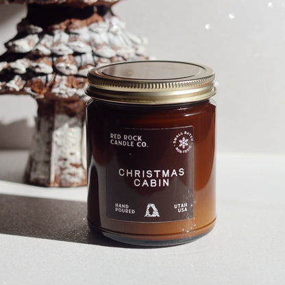 Jar of 'Christmas Cabin' candle from Red Rock Candle Co. with a decorative Christmas tree in the background.
